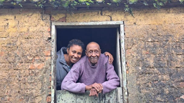 Mulher negra abraça um senhor negro na porta semiaberta de um casa com tijolos à vista.