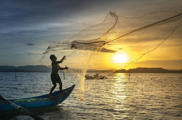 Imagem mostra pescador em cima de um barco e atirando a rede na água. No fundo aparece o sol se pondo.